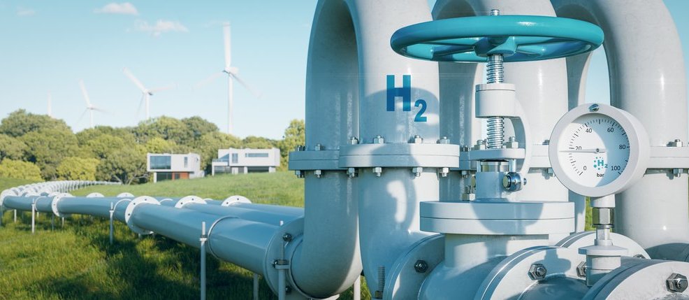 Das Bild zeigt ein Wasserstoff-Industrieventil mit Druckanzeige, beschriftet mit „H₂“. Im Hintergrund sind Windkraftanlagen sichtbar, die die Verbindung zwischen erneuerbaren Energien und Wasserstofftechnologien symbolisieren. Diese Darstellung verdeutlicht die Rolle von grünem Wasserstoff und erneuerbarem Gas als tragende Säule der Energiewende.