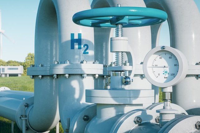Das Bild zeigt ein Wasserstoff-Industrieventil mit Druckanzeige, beschriftet mit „H₂“. Im Hintergrund sind Windkraftanlagen sichtbar, die die Verbindung zwischen erneuerbaren Energien und Wasserstofftechnologien symbolisieren. Diese Darstellung verdeutlicht die Rolle von grünem Wasserstoff und erneuerbarem Gas als tragende Säule der Energiewende.