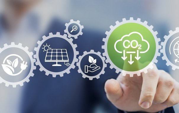 Das Bild zeigt eine visuelle Darstellung der Sektorenkopplung: Zahnräder symbolisieren die Verzahnung verschiedener Energiesektoren wie Strom, Wärme, Gas und Mobilität. Im Vordergrund hebt ein grünes Zahnrad mit dem CO₂-Symbol den Fokus auf CO₂-Reduktion hervor. Weitere Piktogramme wie Solarmodule, Pflanzen und globale Vernetzung verdeutlichen die Bedeutung erneuerbarer Energien und nachhaltiger Lösungen.