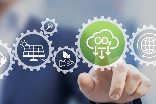 Das Bild zeigt eine visuelle Darstellung der Sektorenkopplung: Zahnräder symbolisieren die Verzahnung verschiedener Energiesektoren wie Strom, Wärme, Gas und Mobilität. Im Vordergrund hebt ein grünes Zahnrad mit dem CO₂-Symbol den Fokus auf CO₂-Reduktion hervor. Weitere Piktogramme wie Solarmodule, Pflanzen und globale Vernetzung verdeutlichen die Bedeutung erneuerbarer Energien und nachhaltiger Lösungen.